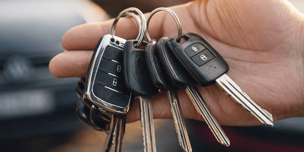Hand hält mehrere Autoschlüssel vor einem Firmenparkplatz – Symbol für mehrere Fahrzeuge und ein zentrales Fahrtenbuch.